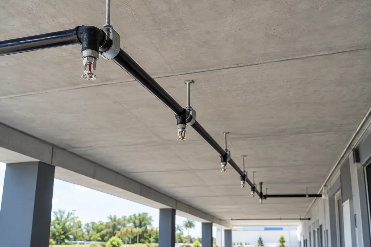 Connected black steel fire sprinkler piping with rigid slab hangers and pendent sprinklers below a commercial concrete canopy in daylight; no people present.