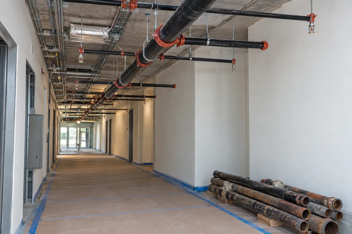 commercial corridor with an open ceiling, new black fire sprinkler mains and branch piping overhead, and removed weathered pipe neatly stacked on protected flooring.