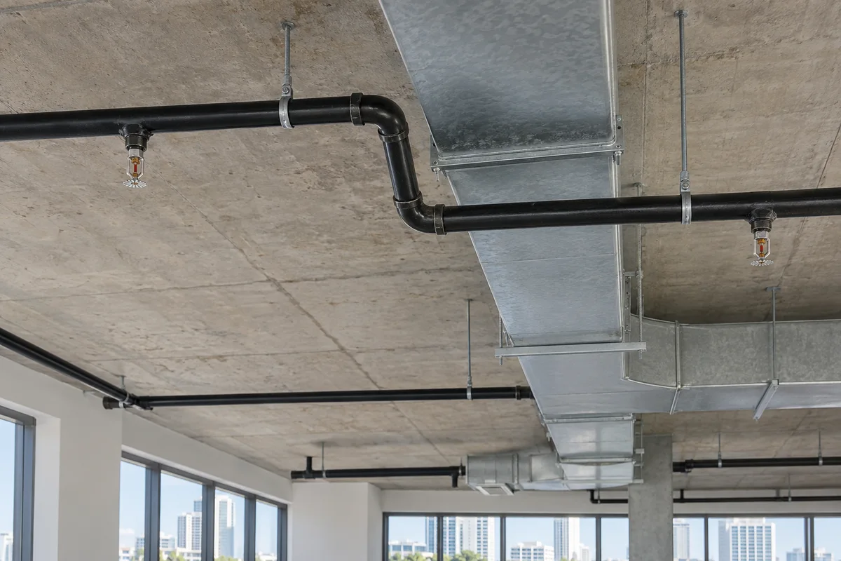 Black fire sprinkler piping offset around an HVAC duct with independent slab hangers and pendent sprinklers below the rerouted branch.