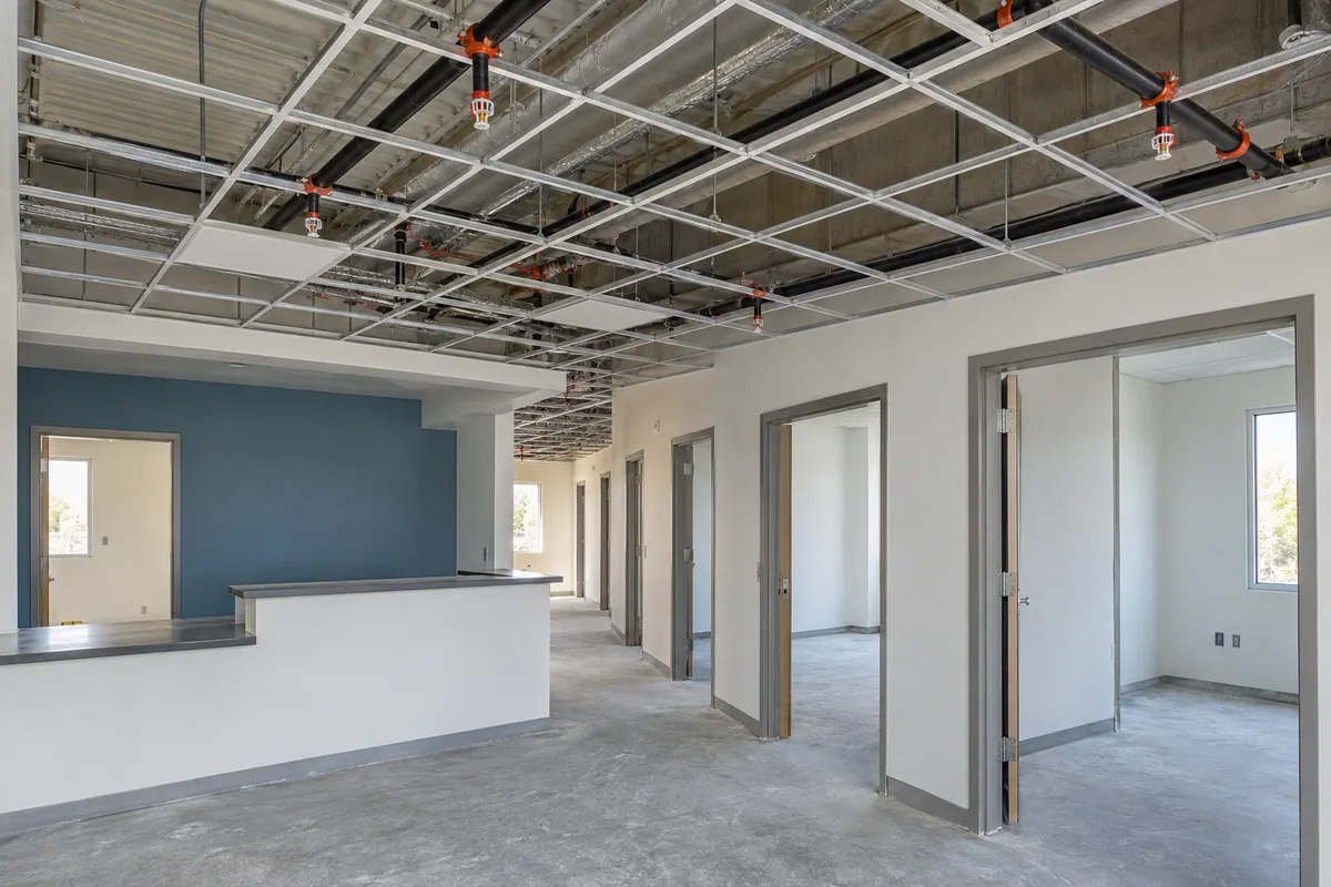 unfinished medical office with reception counter and exam-room corridor, overhead black fire sprinkler piping with pendant sprinkler heads, partial ceiling grid and bare concrete floor.