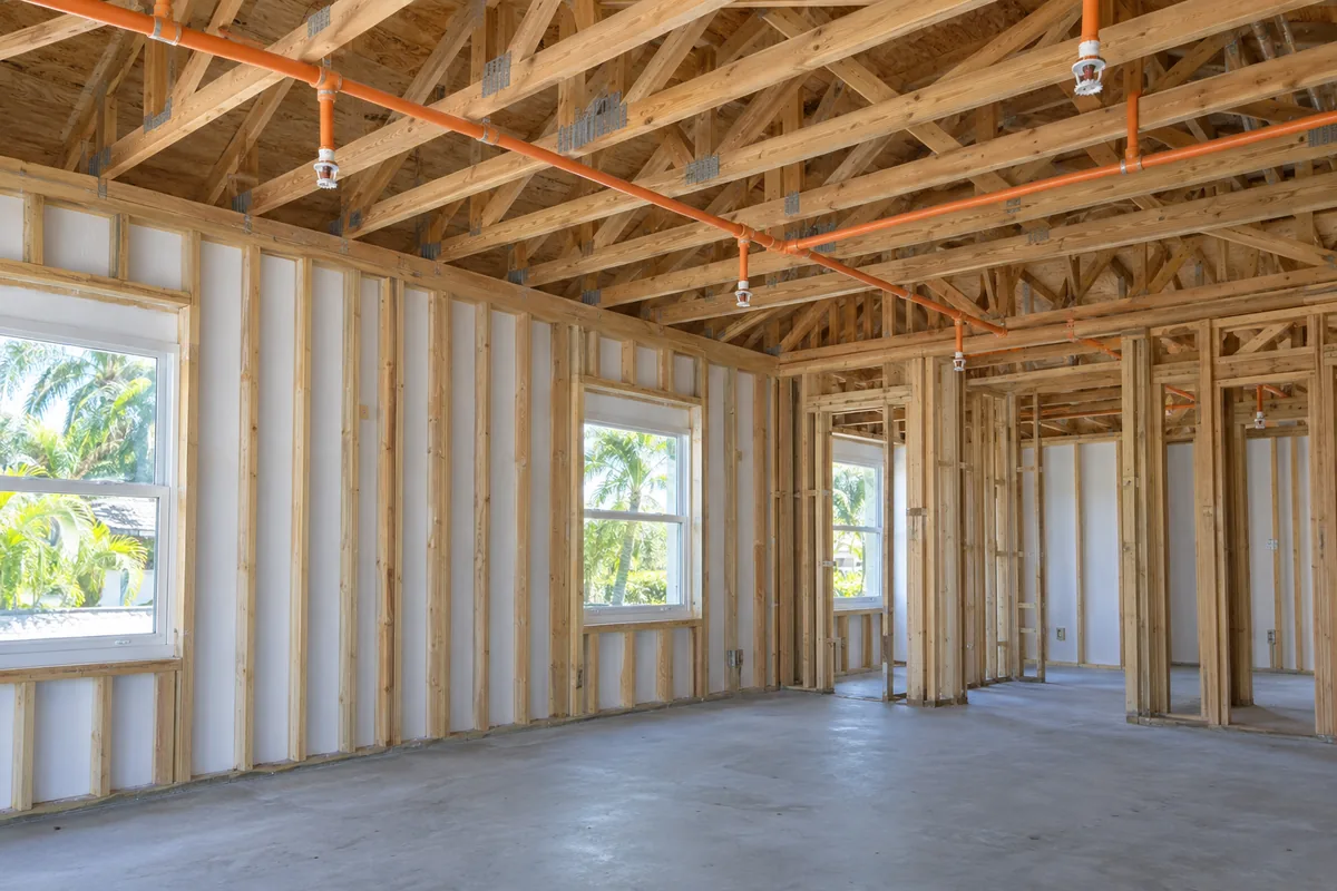 unfinished single-family home framing with wood roof trusses, orange CPVC fire sprinkler branch piping with pendent sprinklers, concrete floor and windows.